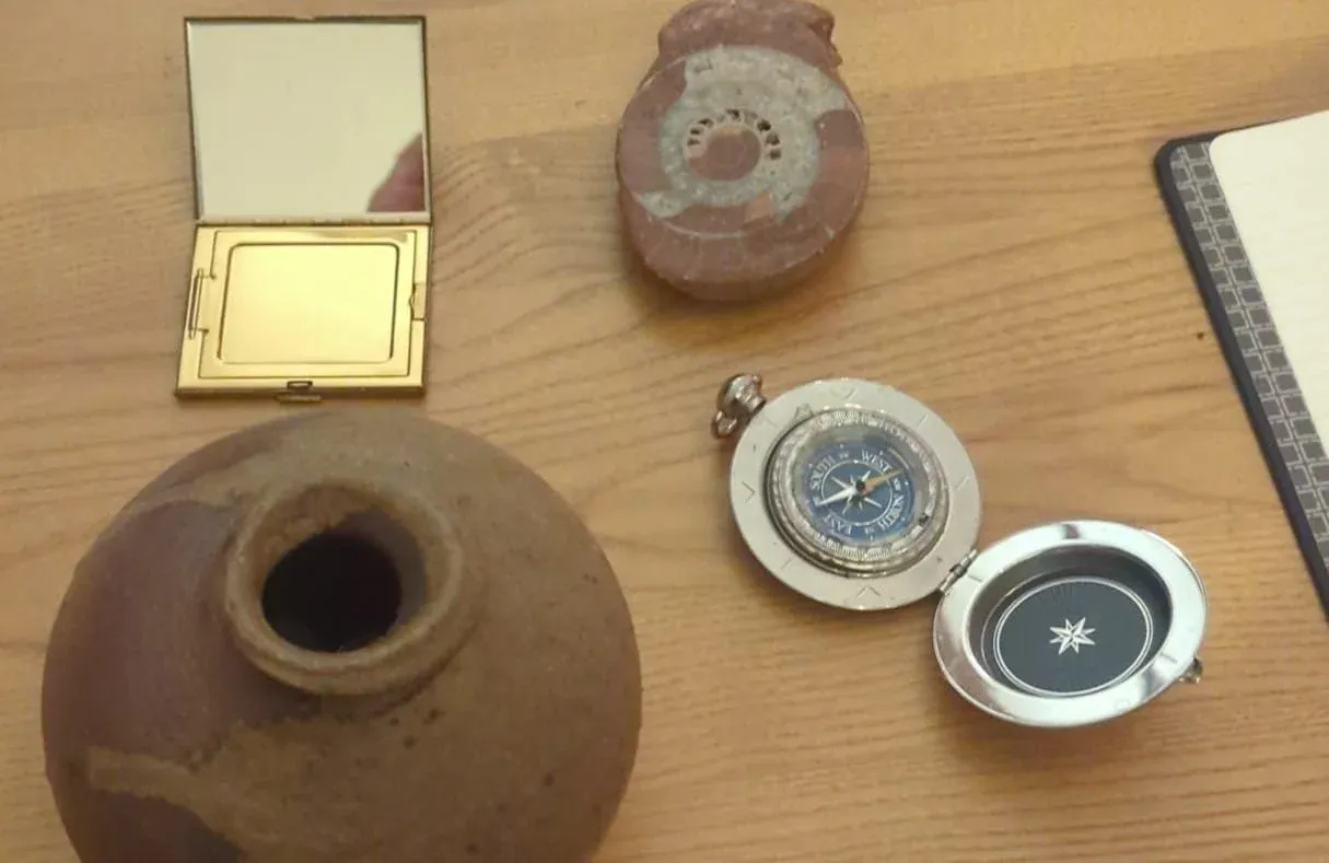 A small earthenware vessel, a brass pocket compass, a folding mirror, and a closed notebook arranged on a wooden table.